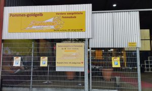 Eingangsbereich der evangelischen Pommesbude „Pommes Goldgelb“ in Verden mit gelben Schildern und Öffnungszeiten.