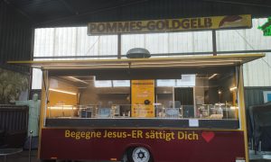 Evangelischer Foodtruck „Pommes Goldgelb“ in Verden mit gelb-rotem Schriftzug und Motto „Begegne Jesus – Er sättigt Dich“.