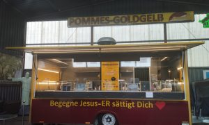 Evangelischer Foodtruck „Pommes Goldgelb“ in Verden mit gelb-rotem Schriftzug und Motto „Begegne Jesus – Er sättigt Dich“.